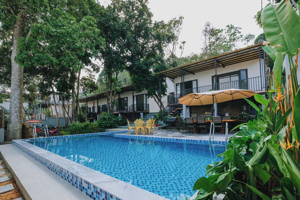 A swimming pool with tiled edges, surrounded by greenery and a building with balconies.