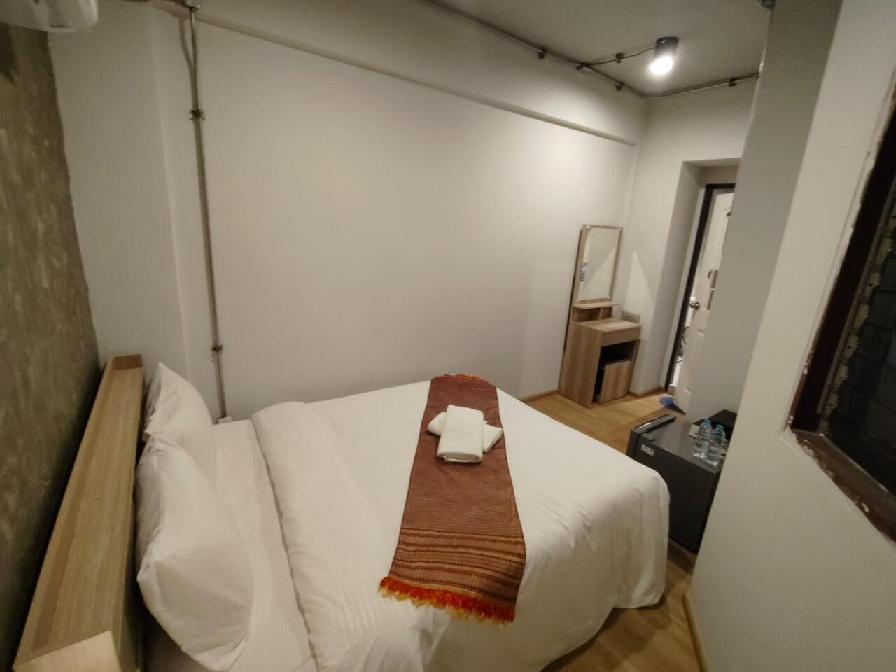 A small, neatly made bed with a white comforter and a brown blanket. There is a small table with a water bottle and a remote control. The room has white walls and a wooden floor.