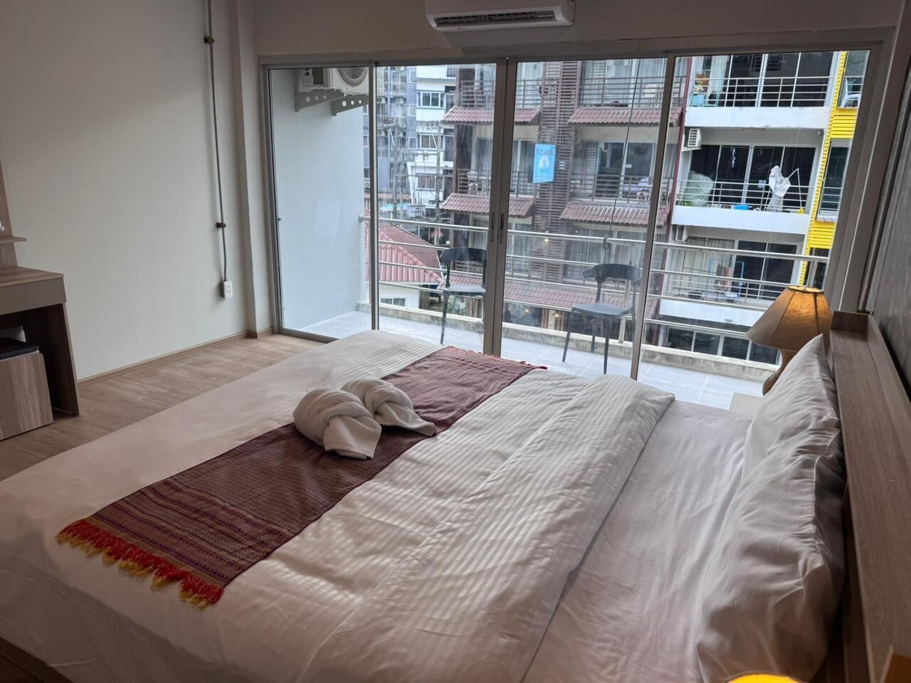 Ein Hotelzimmer mit einem großen Bett, Blick auf eine Stadtlandschaft durch raumhohe Fenster und einem Balkon mit Geländer.
