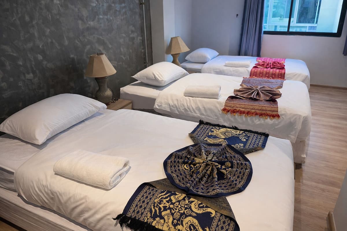 A hotel room with two beds, each with white linens and multiple pillows. There are decorative throw pillows and a blanket on the beds. The room has a modern design with a gray accent wall and a window with curtains.
