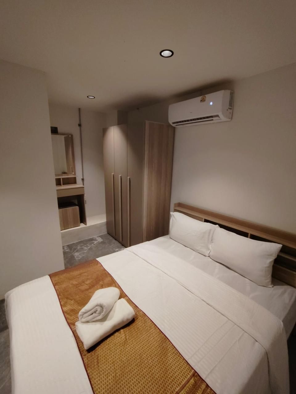 A hotel room with a bed, a wooden wardrobe, a mirror, and an air conditioning unit.