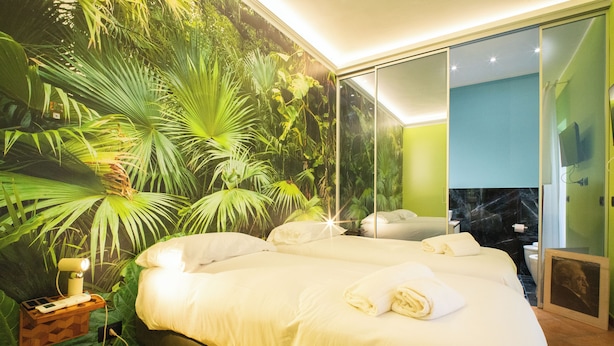 Un dormitorio con una cama grande, una mesita de noche con lámpara y una pared decorada con un mural de plantas tropicales.
