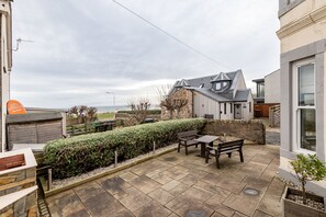 A patio area with stone flooring, wooden benches, and a hedge. - St Albans Lower | Elie (Elie)