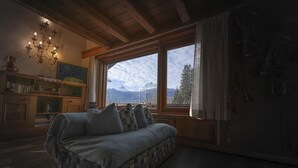 A cozy room with a checkered sofa, a wooden ceiling, a large window with a mountain view, and a wall-mounted shelf with books and decorative items. - Casa Funivia 2, close to the cable car and the center (Cortina d´Ampezzo)