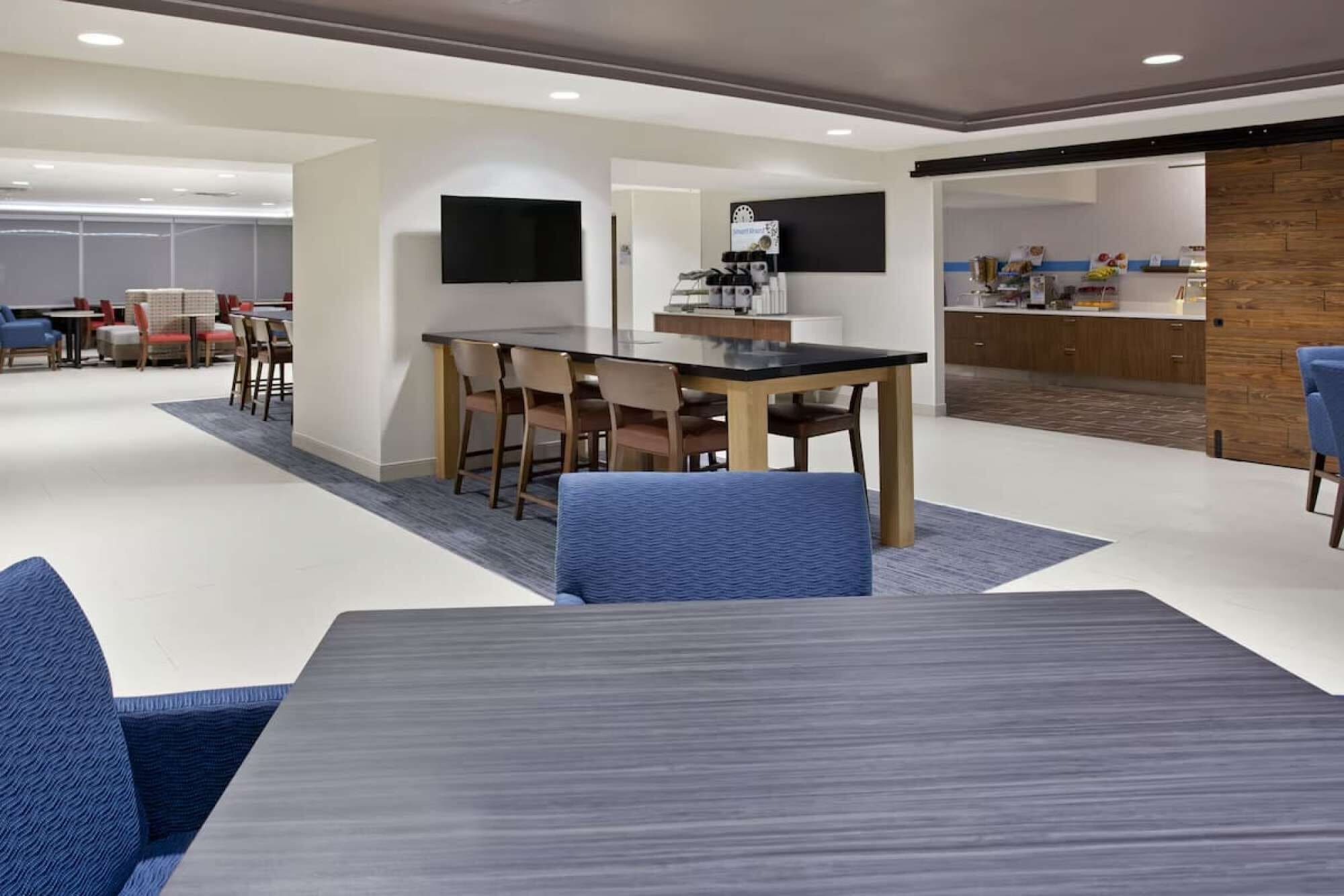 A modern hotel lobby with a dining area, a kitchen, and a seating area with blue chairs and a table.