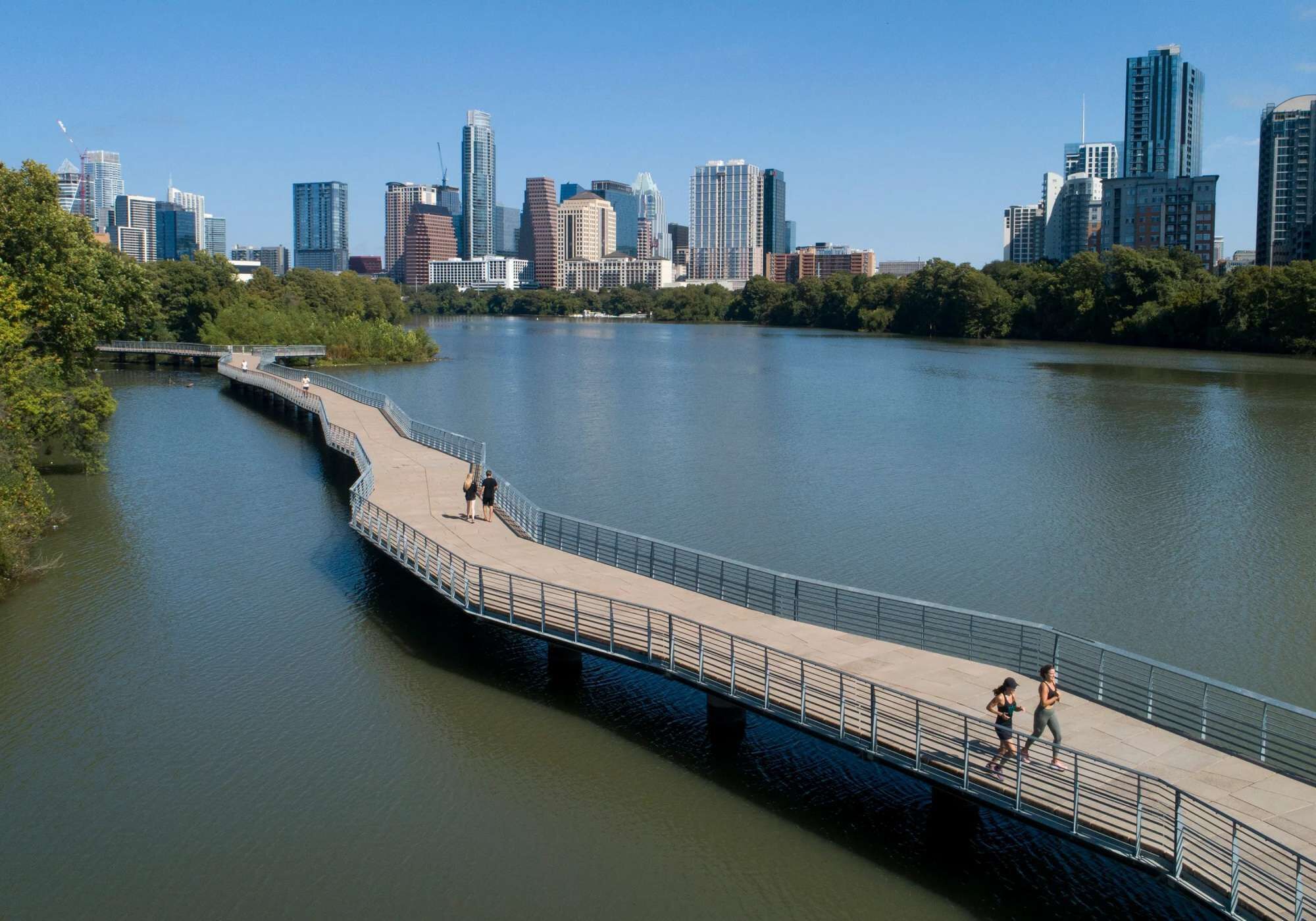 A riverside walkway with a metal railing, leading to a city skyline in the background.