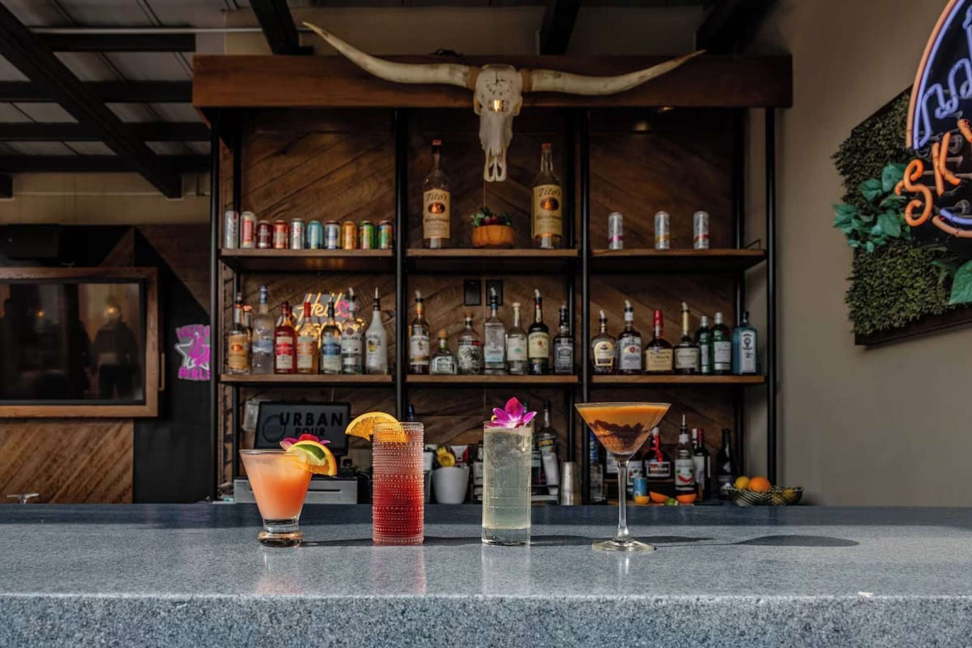 Un coin bar avec une variété de bouteilles d’alcool, une décoration en forme de crâne et trois cocktails différents présentés sur le comptoir.