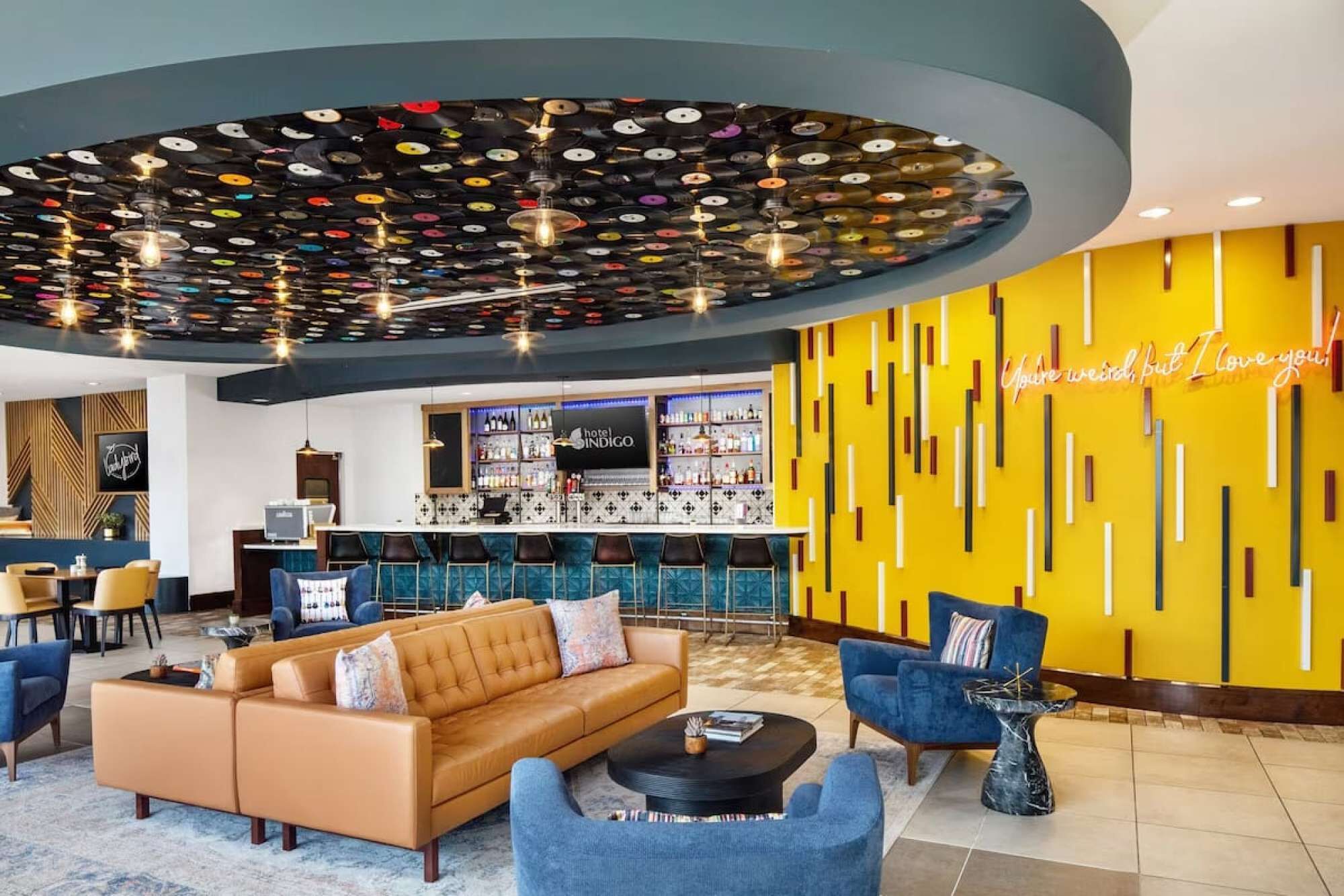 Um saguão de hotel com projeto de teto circular, área de bar e espaço de estar com parede de destaque amarela.