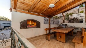 Un área de comedor al aire libre rústica, con una mesa y bancos de madera, una chimenea y una pérgola.