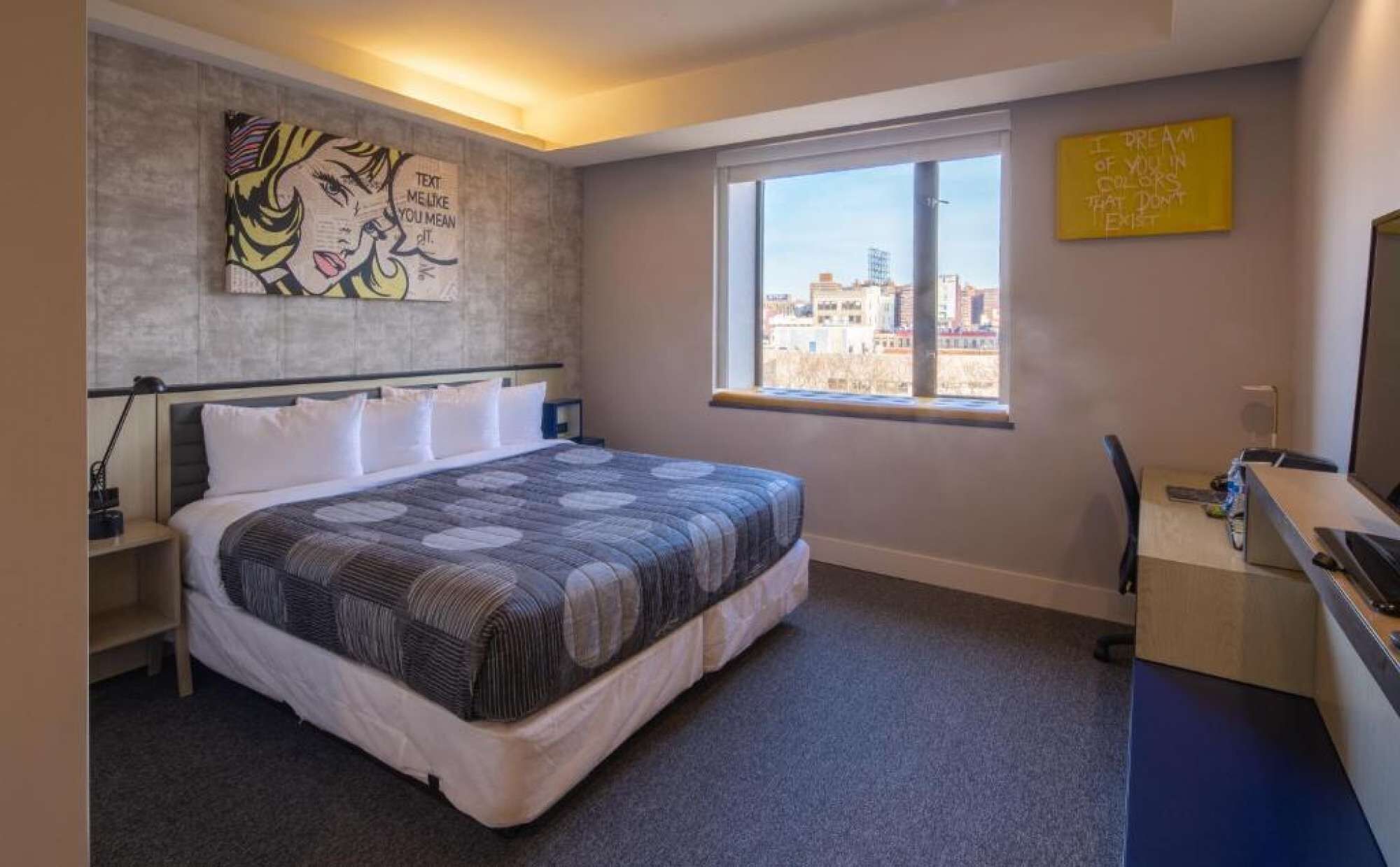 A modern bedroom with a large bed, a desk, and a window with a city view.