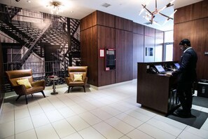 A lobby with a staircase, two leather armchairs, a reception desk with laptops, and a fire safety panel. - Steps From The Lower East Side | Sightseeing (New York)