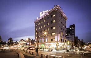 A multi-story hotel with a prominent sign at the top, multiple ground-floor shops, and a well-lit street scene. - Steps From The Lower East Side | Sightseeing (New York)