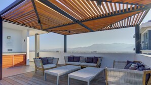 A rooftop terrace with wooden flooring, a covered seating area with cushions, and a view of distant mountains. - Stunning Ocean+Marina Views Rooftop Infinity Pool (Puerto Vallarta)