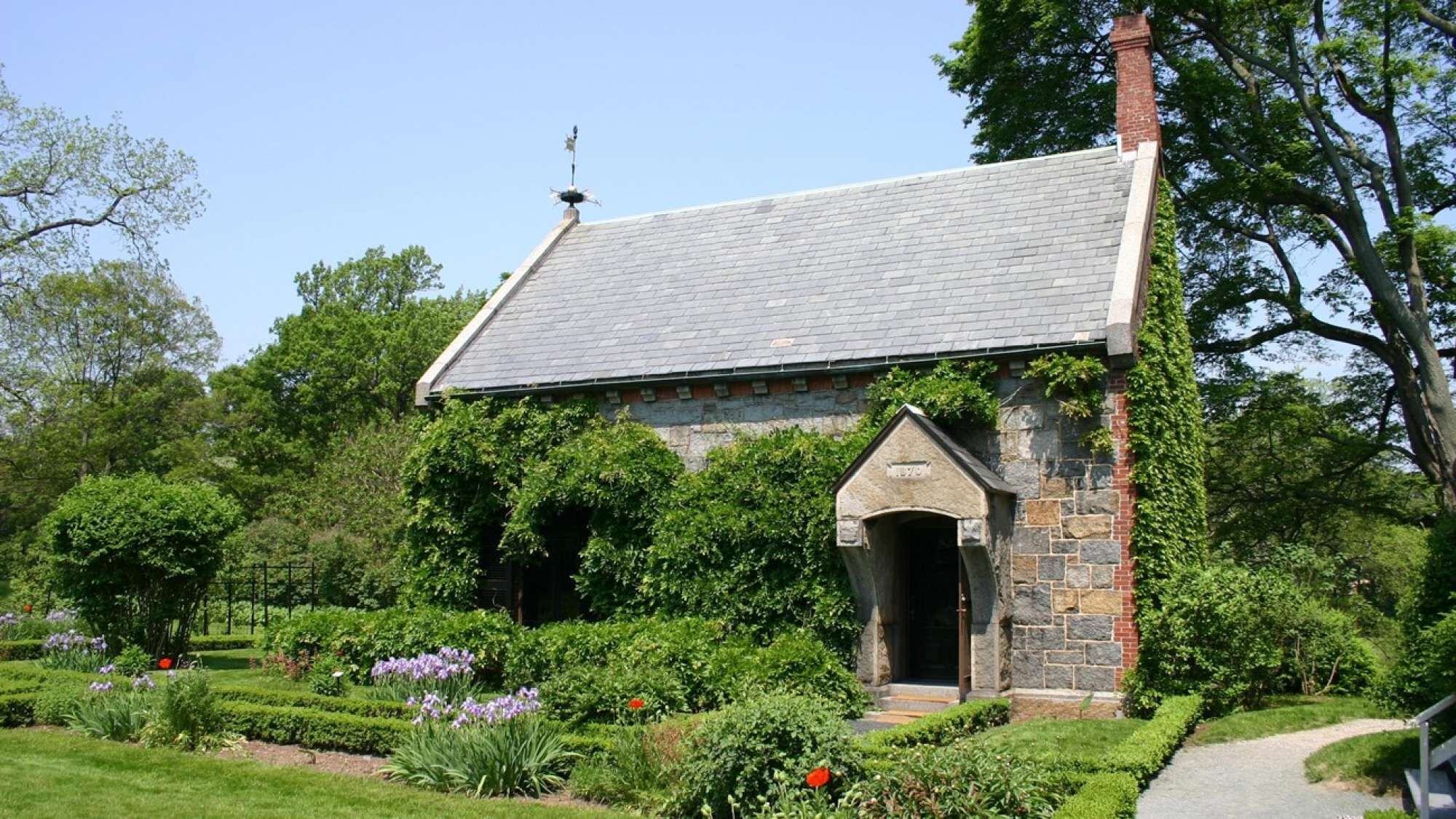 A stone building with a sloped roof, surrounded by lush greenery and flowering plants.