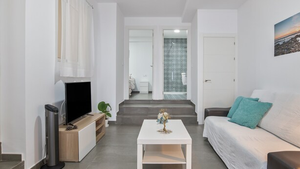 Een moderne woonkamer met een flatscreen televisie, een witte salontafel, een bank met blauwgroene kussens en een trap naar een badkamer.