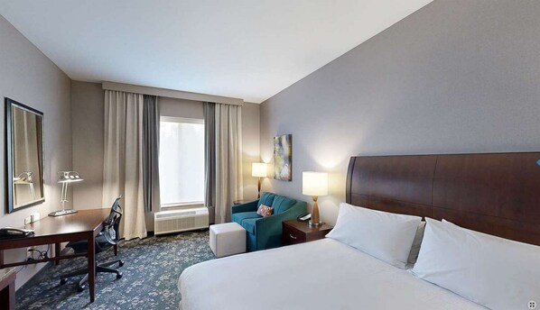 A hotel room with a large bed, a desk with a chair, a small table with a lamp, a window with curtains, and a painting on the wall.