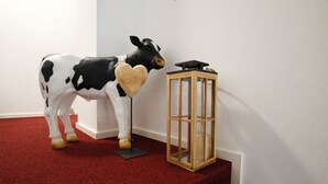 Una estatua de vaca con un objeto en forma de corazón en la boca, ubicada junto a un farol de madera sobre un piso alfombrado de rojo.