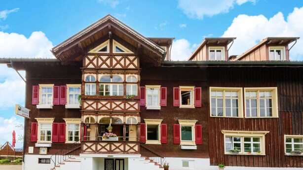 Un edificio de estilo chalete tradicional con techo inclinado, varias ventanas con contraventanas y un balcón con macetas colgantes de flores.