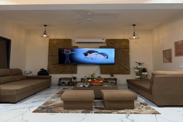 Ein Wohnzimmer mit einem wandmontierten Flachbildfernseher, einer Couch, zwei Hocker, einem Couchtisch mit Früchten und einer dekorativen Wandverkleidung.