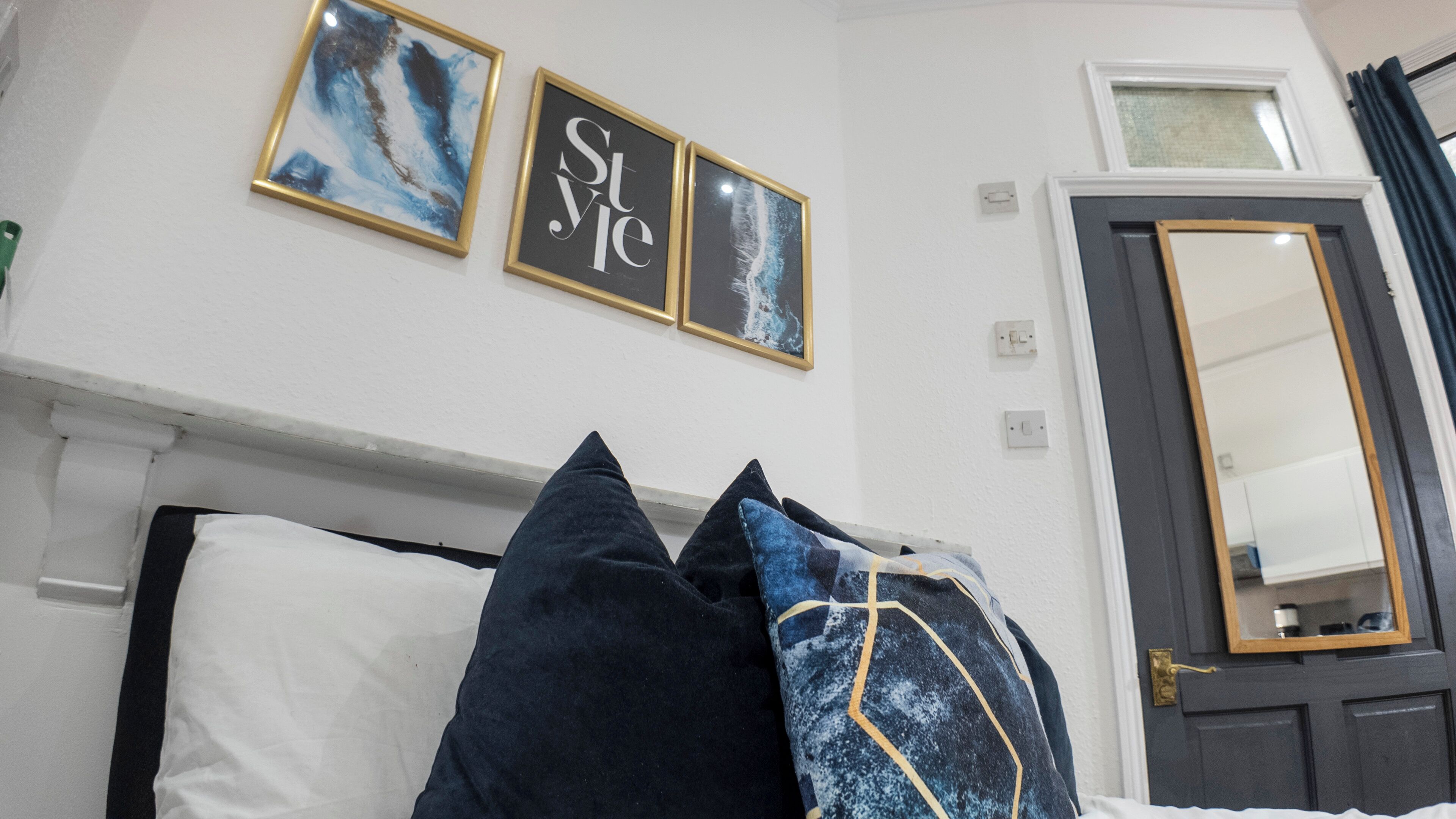 A bedroom with a bed, pillows, a wall-mounted mirror, and two framed artworks.
