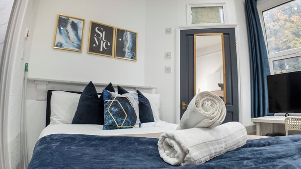A neatly made bed with a blue blanket, a white towel, and decorative pillows. There is a framed artwork above the bed and a television on a stand to the right. - Spacious Charming Studio in Birmingham (Birmingham)