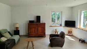 A living room with a sofa, a chair, a wooden cabinet, a TV, and a window with a view of greenery. - Le nouvel Érable, country house a haven of peace and greenery (Chamant)