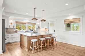 Une cuisine moderne avec des meubles blancs, un grand îlot central doté de tabourets, un éclairage suspendu et une vue sur la verdure à l’extérieur.