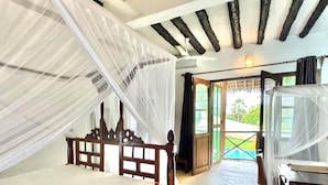 A bedroom with a wooden bed, a desk, and a balcony with a view of greenery. - Pangachumvi Beach Resort (Matemwe)