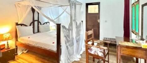 A room with a wooden bed under a canopy, a chair, a desk with a mirror, and a wooden floor.