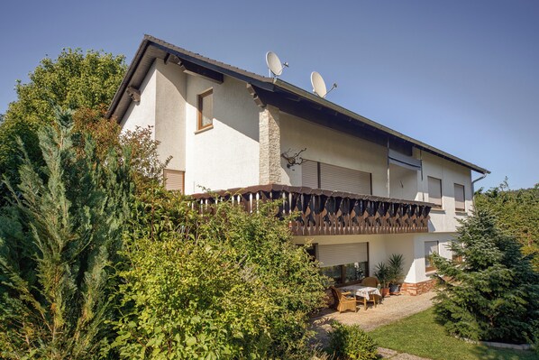 Une maison à deux étages dotée d’un balcon, d’antennes paraboliques et d’un jardin aménagé avec du mobilier d’extérieur.
