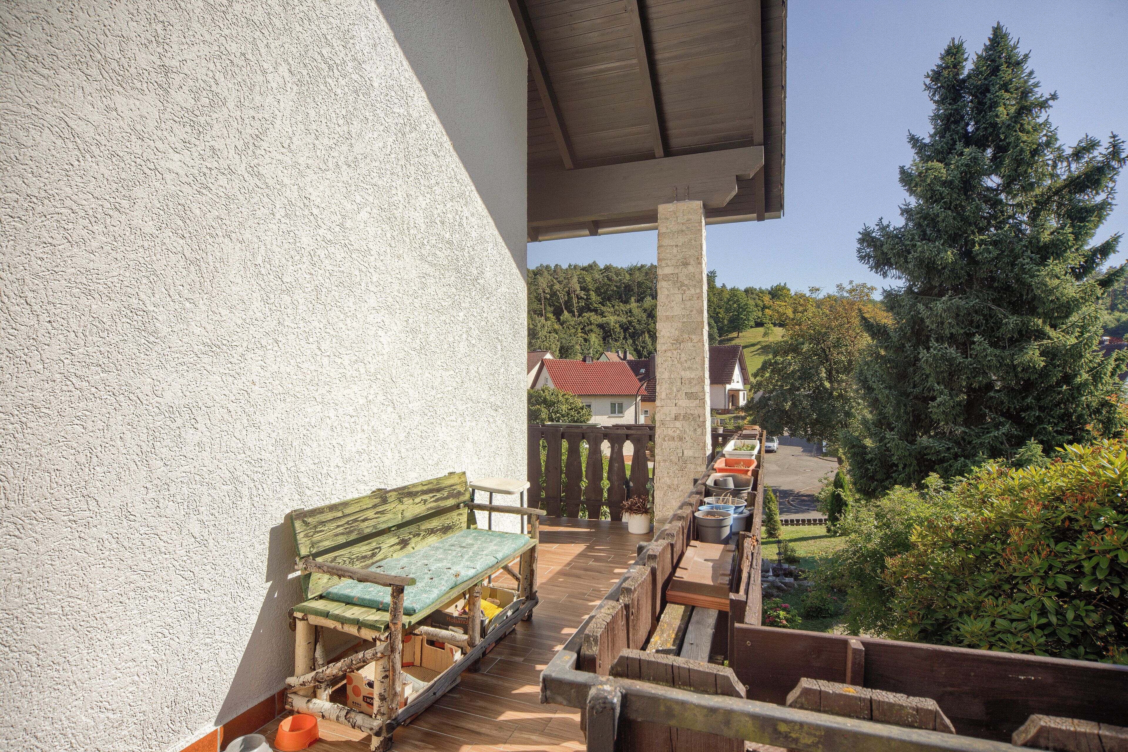 Un balcone con una panchina in legno, una pianta in vaso e vista su una zona residenziale con case e alberi.