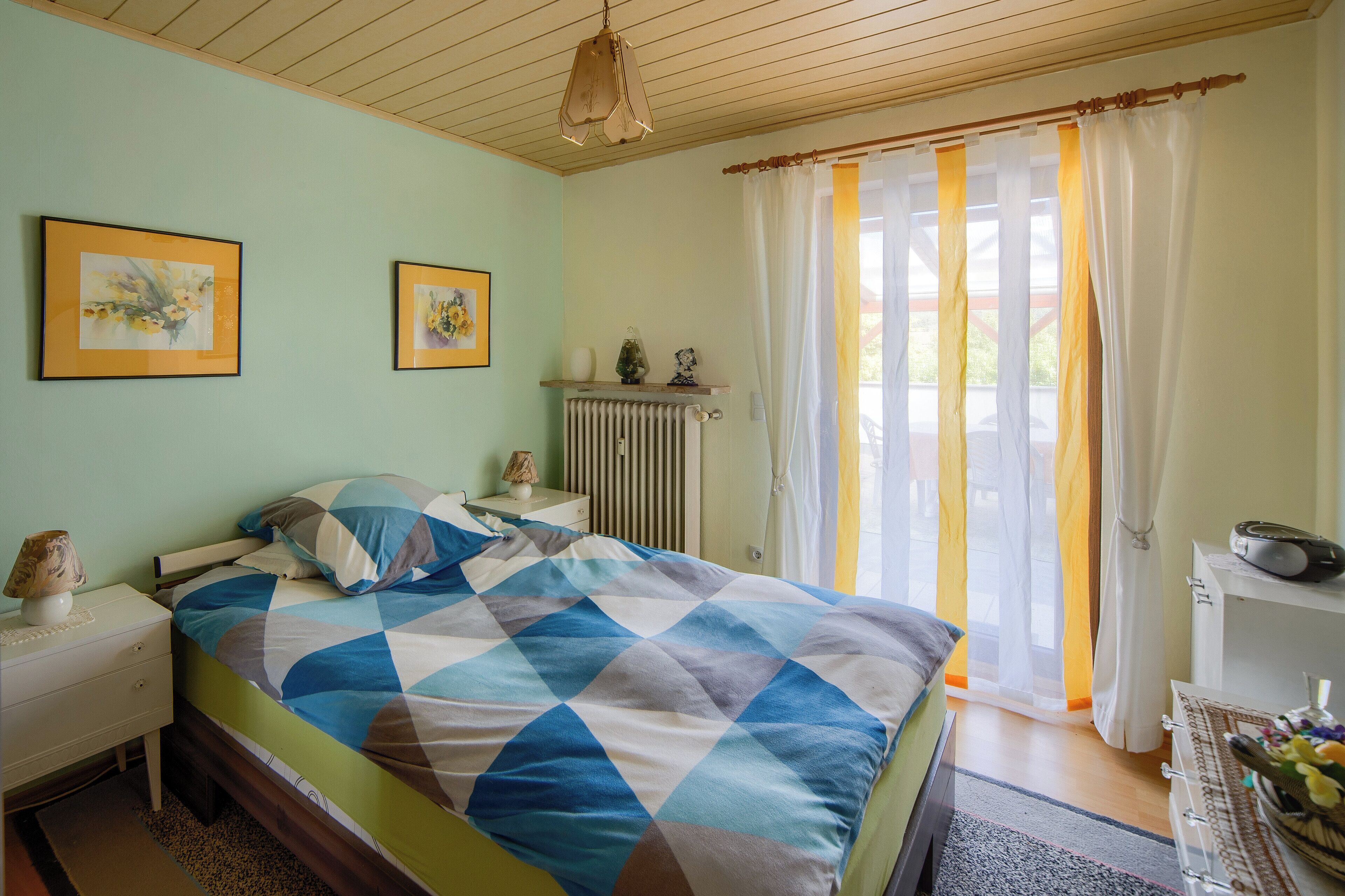 Une chambre à coucher comprenant un lit, une table de chevet, une commode, un radiateur et une fenêtre avec des rideaux.