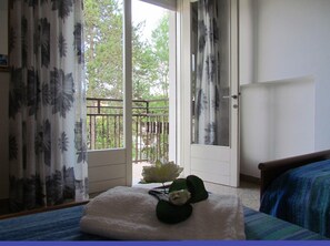 Ein Schlafzimmer mit Bett, Fenster mit Vorhängen, Balkon mit Geländer und einer Tür.