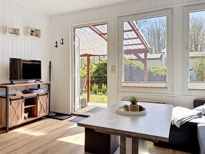 A room with a TV, wooden furniture, a table with a plant, and large windows.