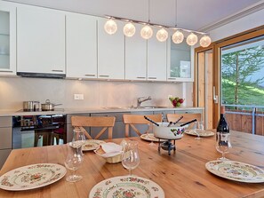 A modern kitchen with white cabinets, a dining table set with floral plates, and a view of greenery outside. - Perrine by Interhome (Saas-Fee)