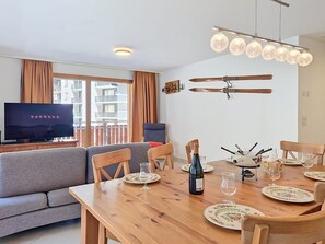 A modern living room with a flat-screen TV, a wooden dining table set for four, and a sofa. - Perrine by Interhome (Saas-Fee)