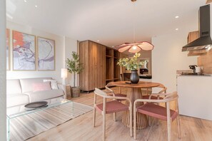 En moderne stue med sofa, sofabord i glas og spiseområde med et rundt bord og stole. Der er også et køkken med træskabe og komfur.