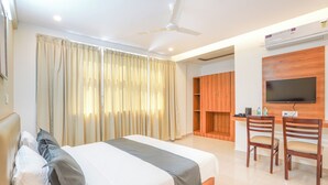 Une chambre d’hôtel avec un lit, des rideaux, un ventilateur de plafond, une télévision et un bureau avec des chaises.