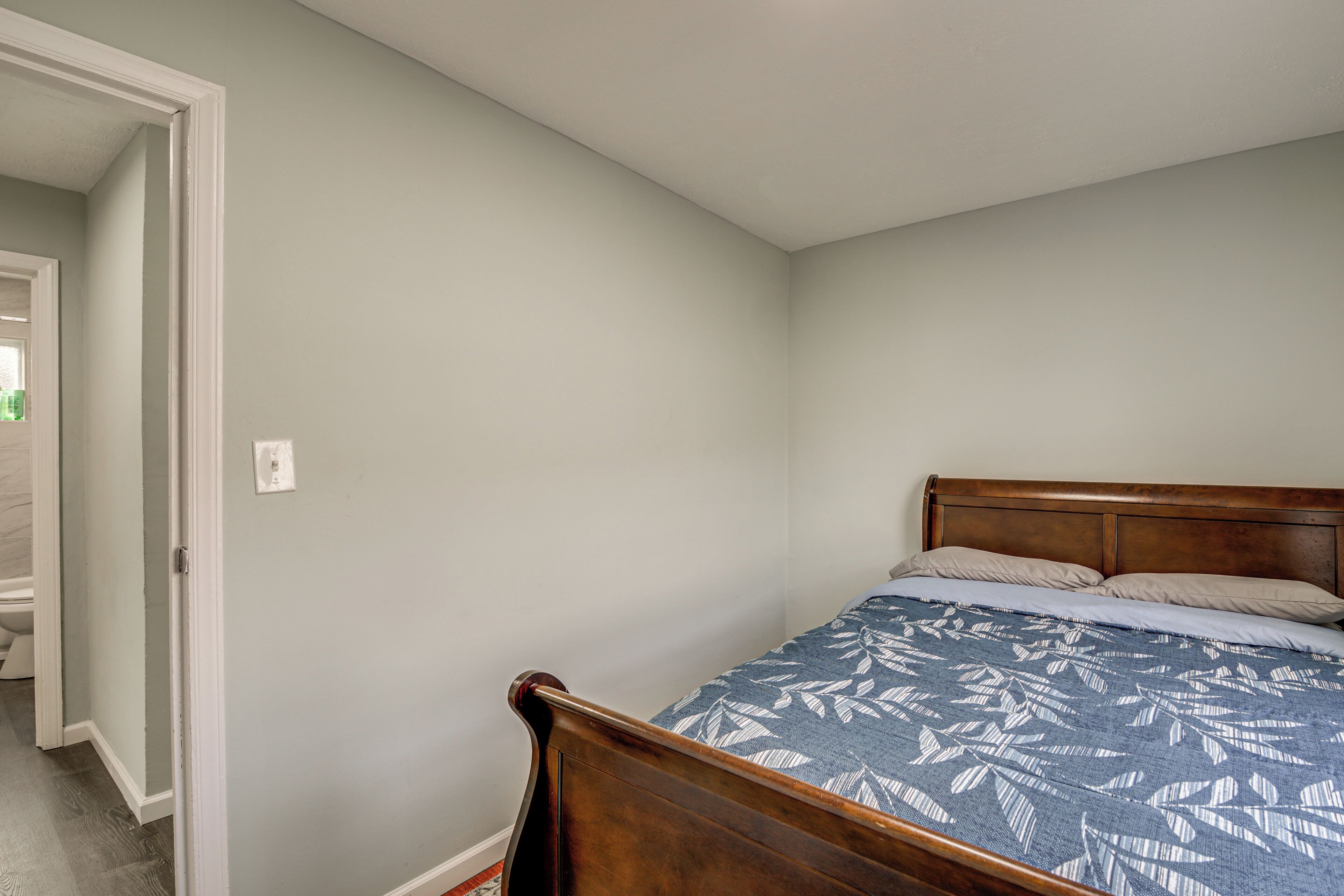 A bedroom with a wooden bed frame, a blue patterned bedspread, and a light-colored wall.