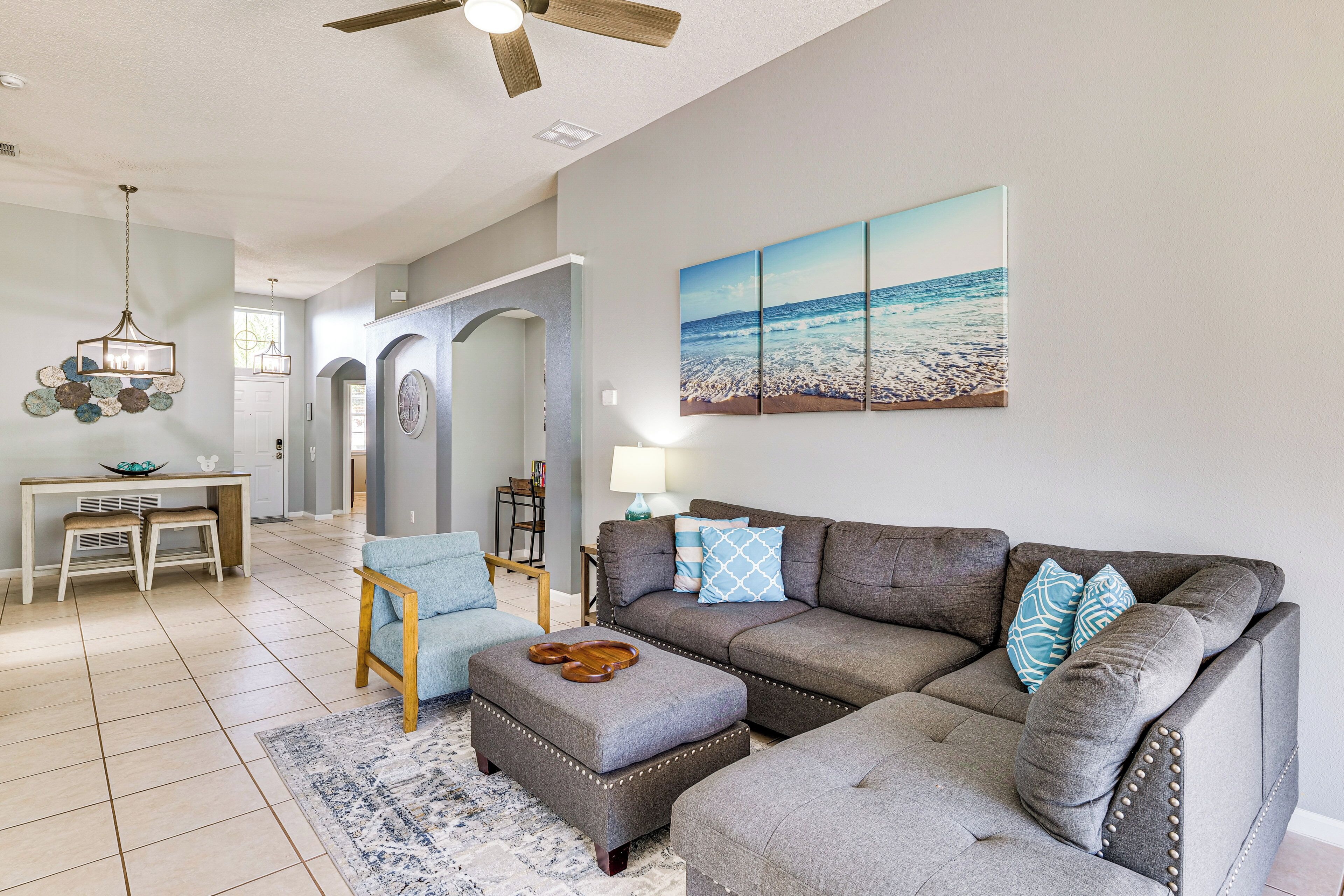 A living room with a gray sectional sofa, a wooden coffee table, and a blue armchair. There is a large beach-themed painting on the wall and a ceiling fan above.