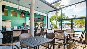 An outdoor seating area with wicker chairs and tables, a pool in the background, and a bar area with a green wall and ceiling fans. - Resort Perks! Family Home 4 Mi to Disney World (Kissimmee)
