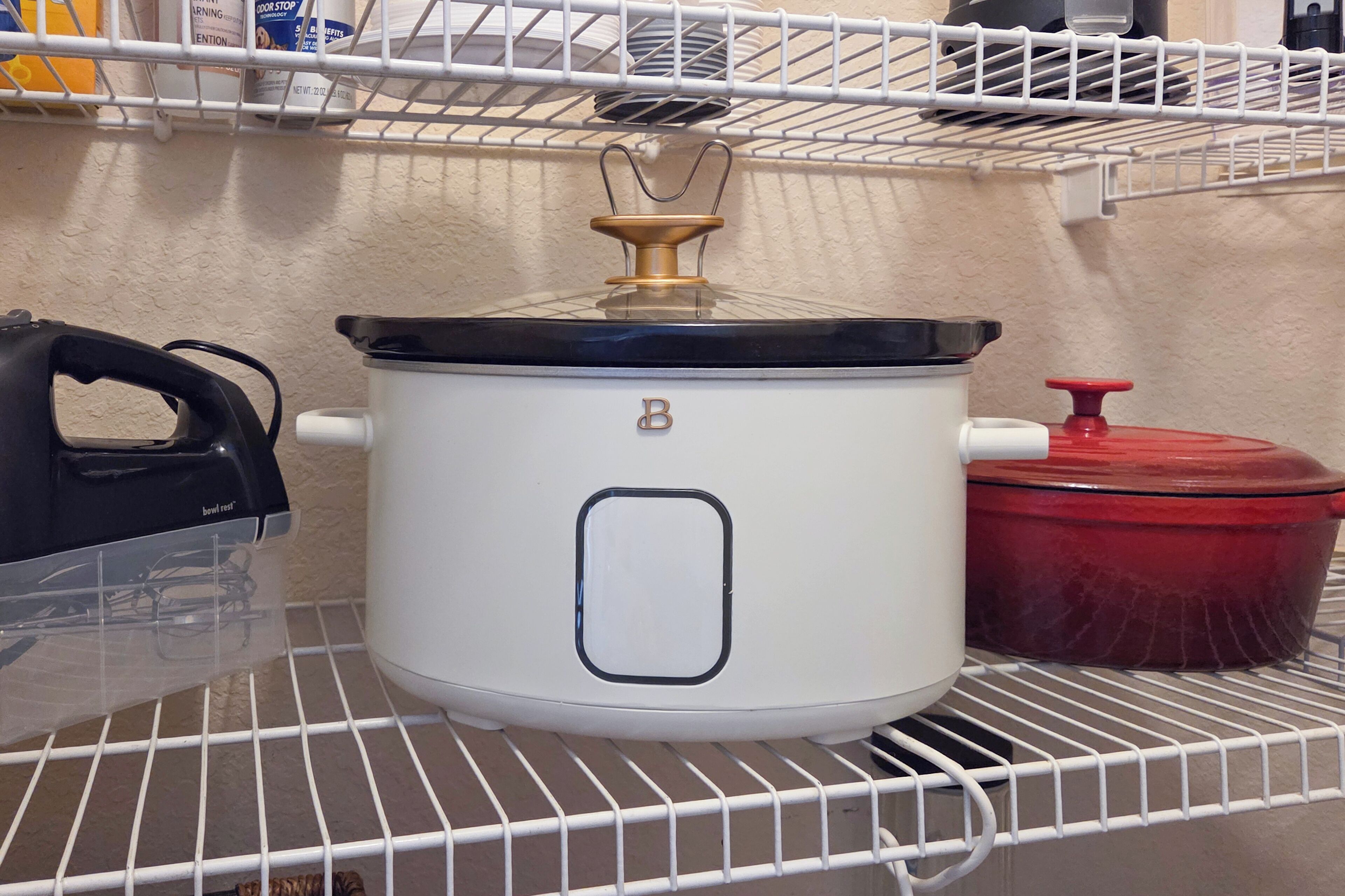 A white slow cooker on a shelf with a red pot and a black appliance on the left.