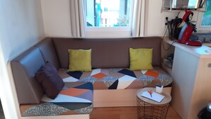 A compact living space with a sofa, cushions, a small round table, and a kitchenette area. - Mobile home 3 bedrooms 6 pers on camping cap soleil Oléron (Saint-Denis-d'Oléron)
