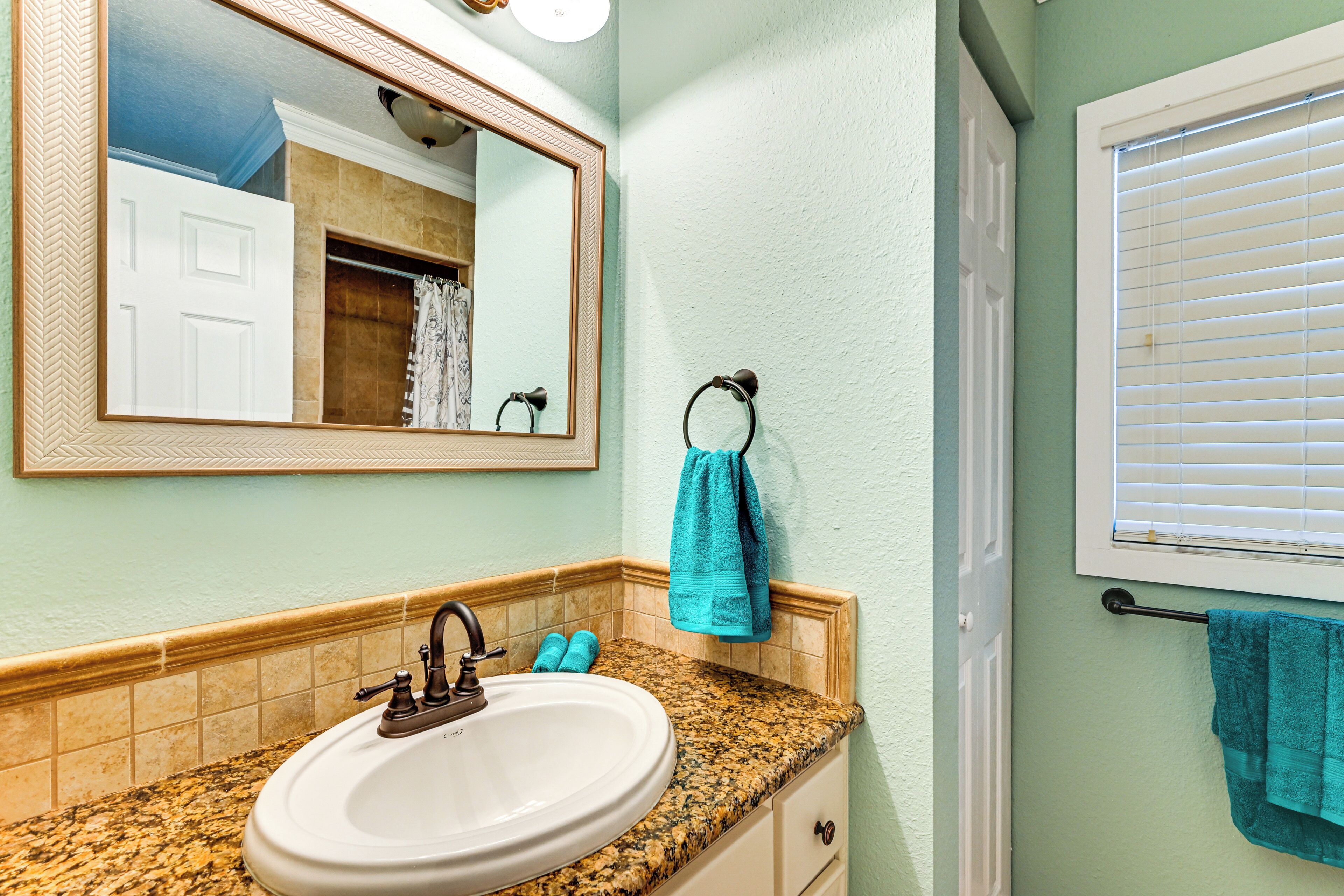 Une salle de bain avec un lavabo doté d’un plan de travail en granit, un miroir, un porte-serviettes et une fenêtre avec des stores.