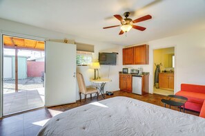 Une chambre d’hôtel de taille réduite, comprenant un lit, un petit bureau avec une lampe, une kitchenette et un ventilateur de plafond.