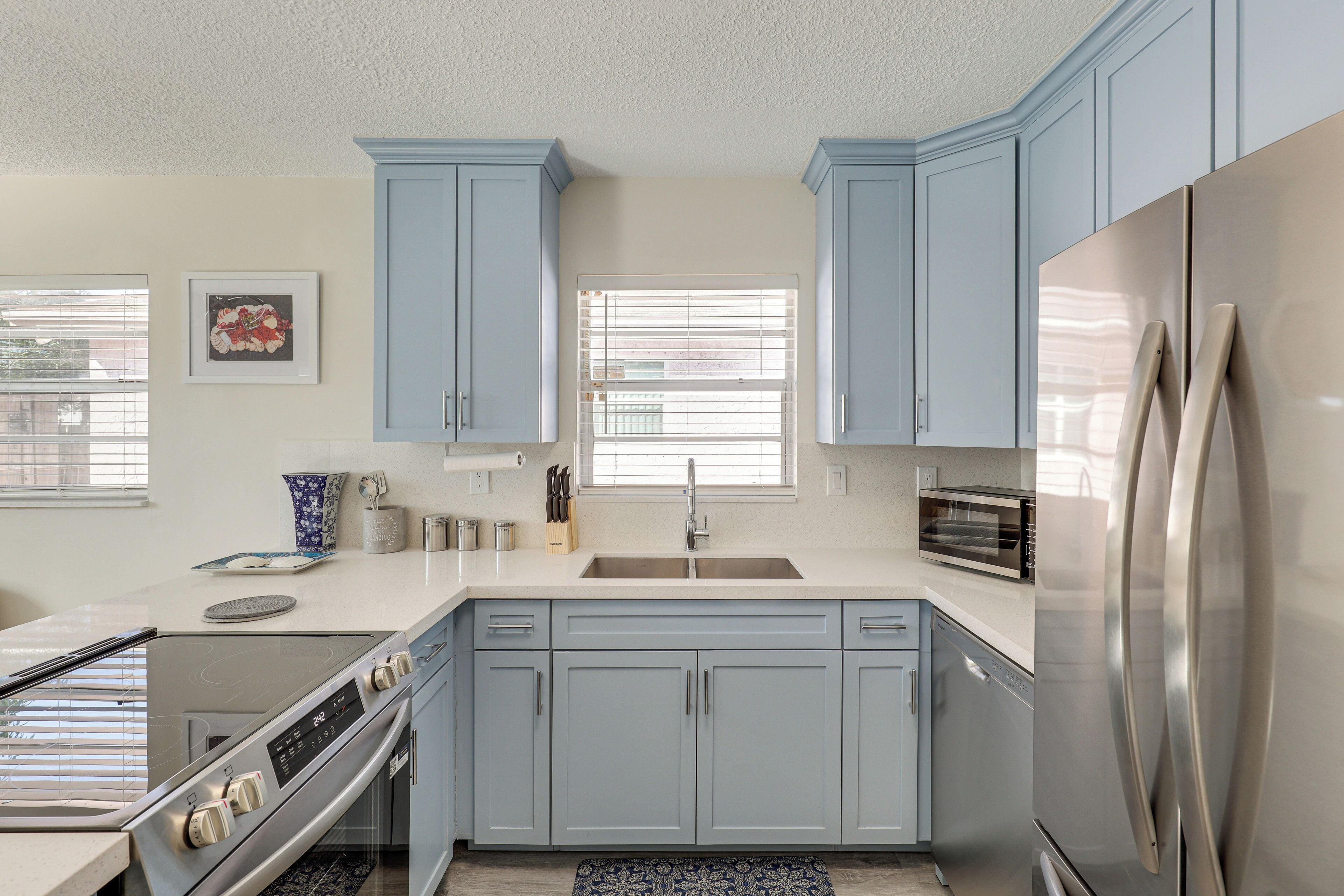 Une cuisine moderne avec des meubles de couleur bleu clair, des appareils en acier inoxydable et une fenêtre équipée de stores.