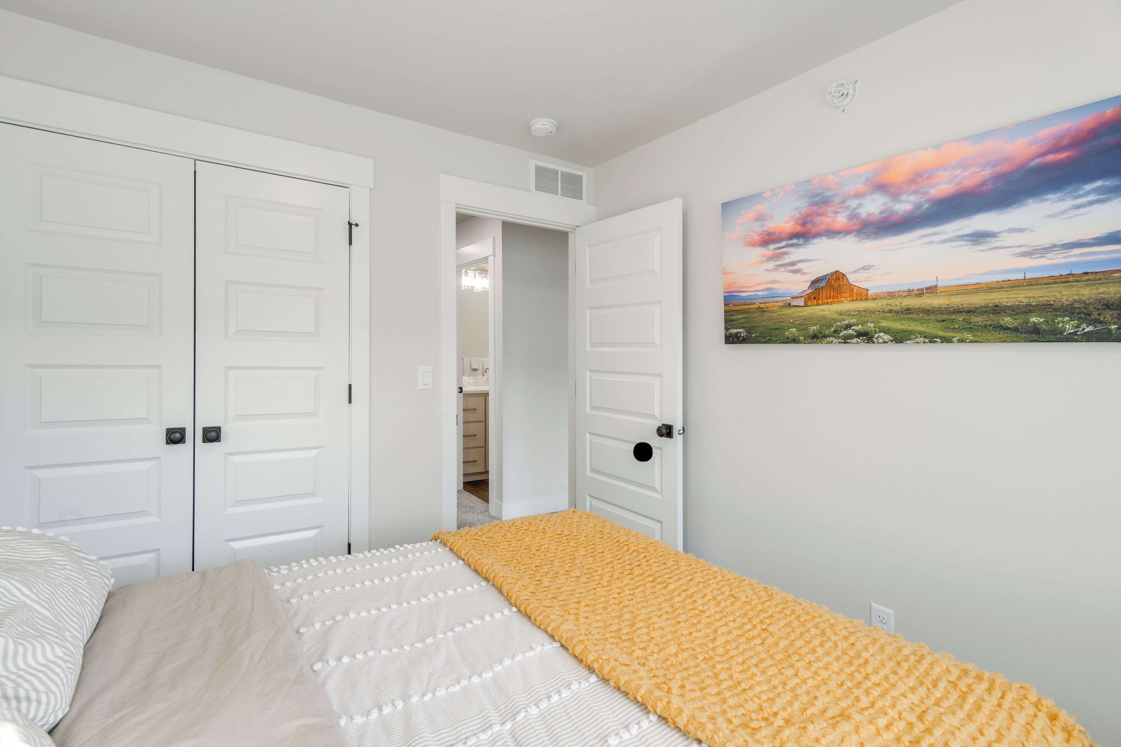 Une chambre à coucher avec un lit, une couverture jaune, des murs blancs et un tableau représentant une grange.