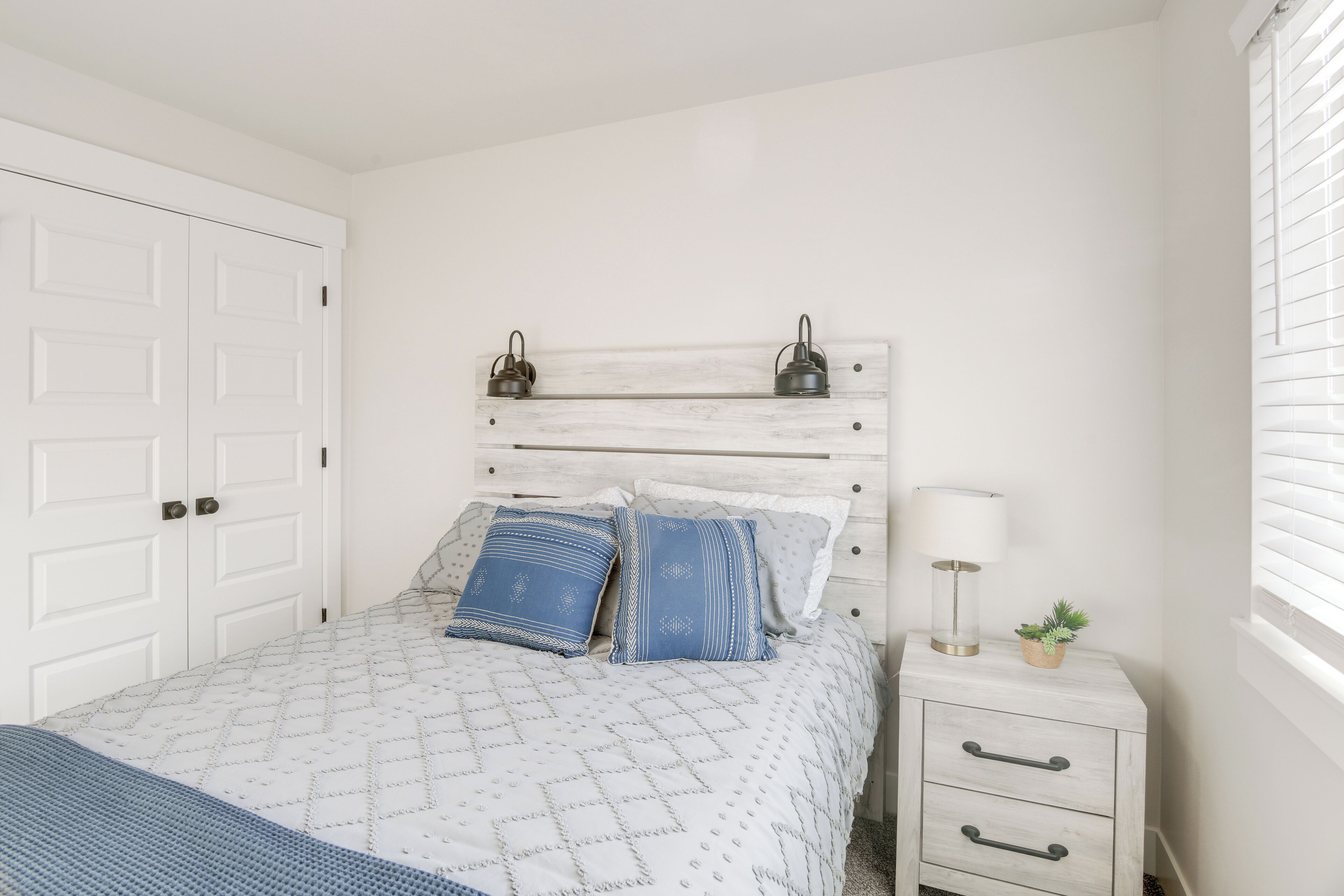 A bedroom with a bed, a nightstand, a lamp, and a window with blinds.
