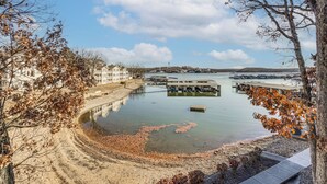 A waterfront view with a sandy beach, a dock, and buildings in the background. - Balcony w/ Lake Views: Waterfront Ozarks Condo! (Osage Beach)