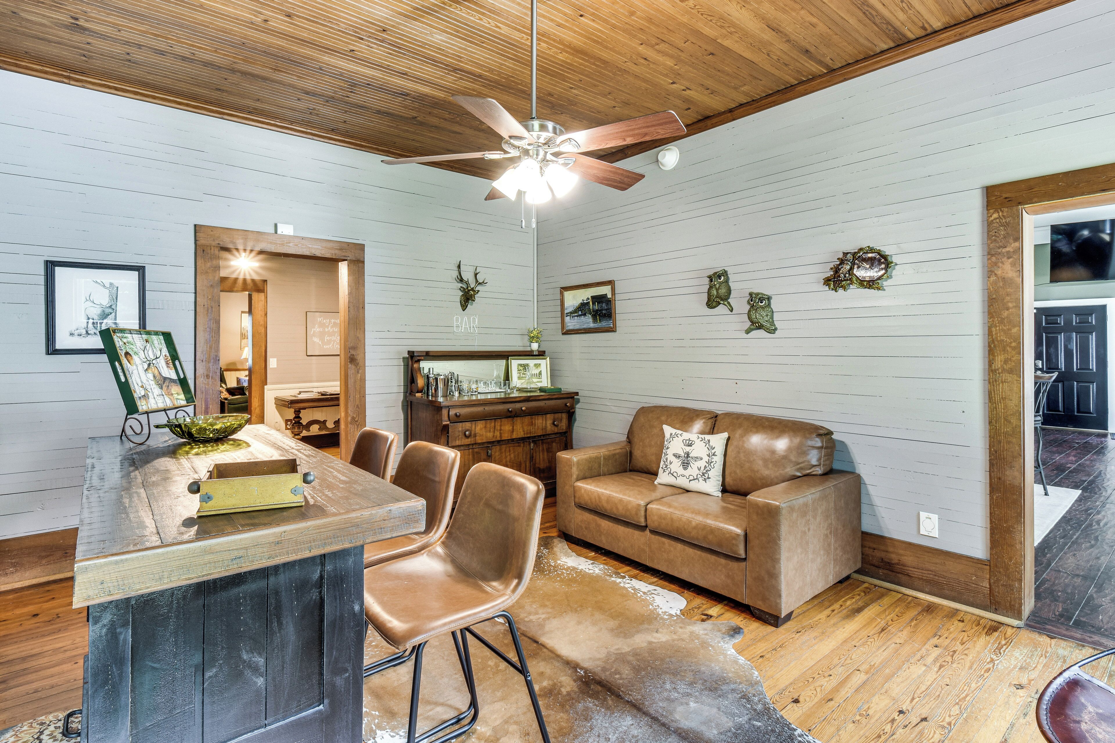 A rustic interior with a wooden ceiling, a ceiling fan, a bar area with stools, a leather sofa, and a cowhide rug.